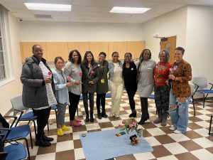 a group of women smile in a circle while holoding flowers, a small cloth with a bouquet of flowers sits in the center of the group.